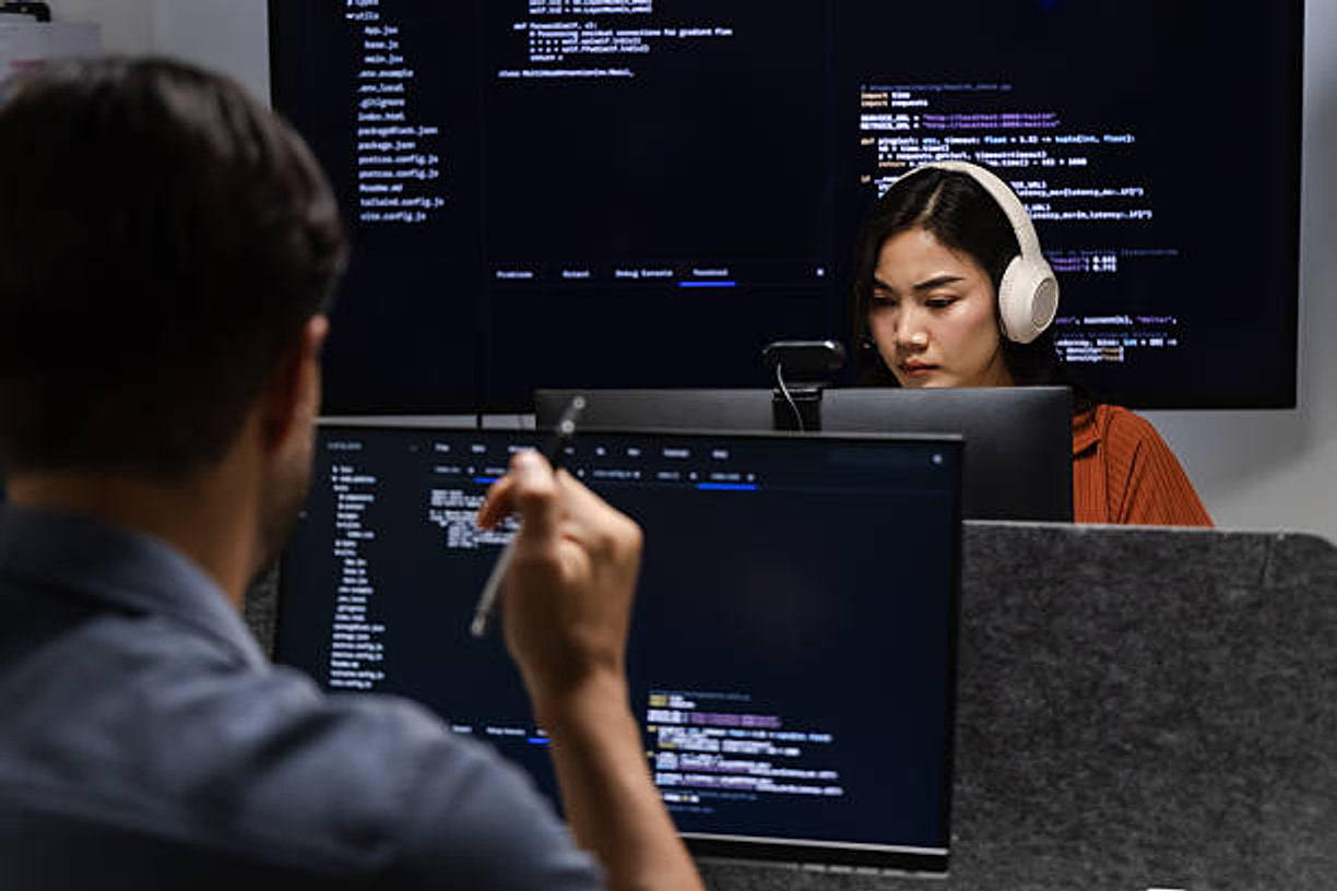 Engineers at their screens, one pointing through code while a colleague works beside them.
