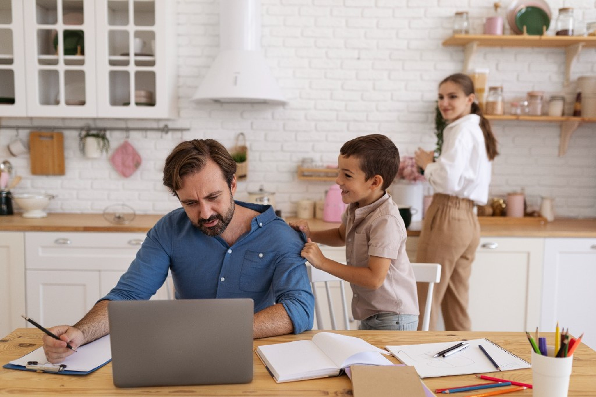A father working at the kitchen table while his son stands beside him and family life continues in the room.