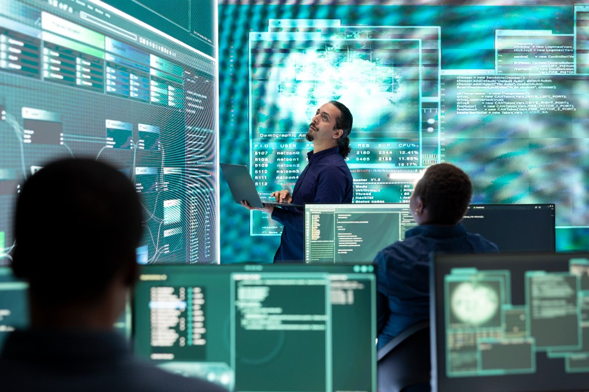 Engineers in a room of large data screens, one standing with a laptop.