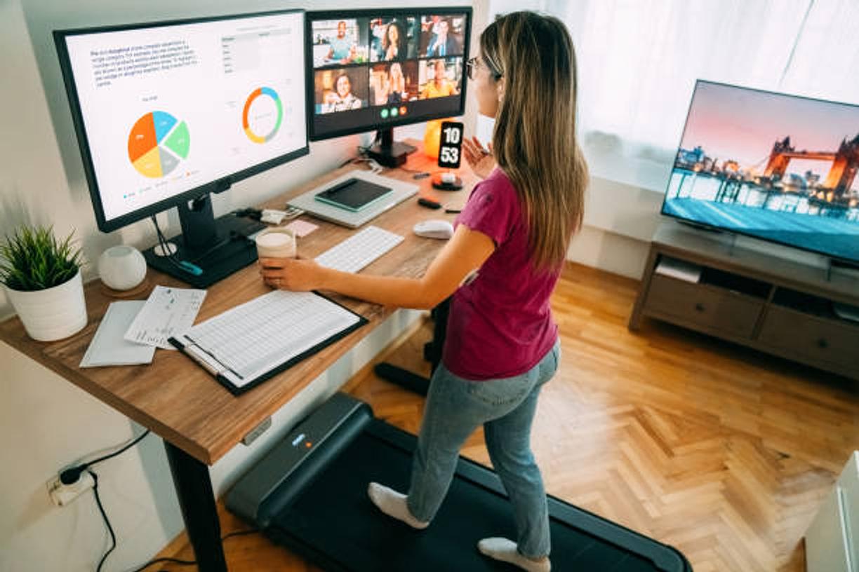 A woman working at a standing desk at home, with a treadmill underfoot and a video call on the second screen.