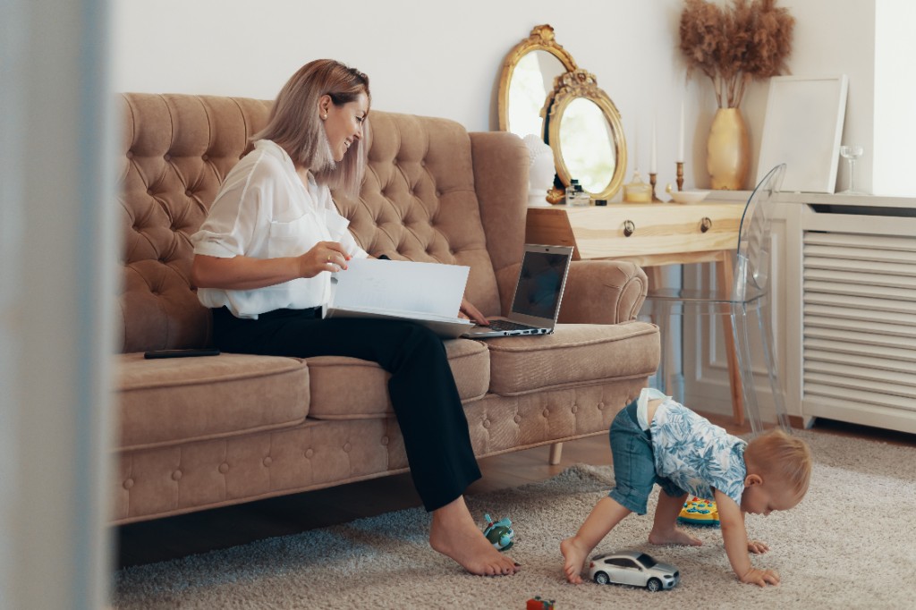 A mother on a sofa with papers and a laptop, while her toddler plays on the rug.