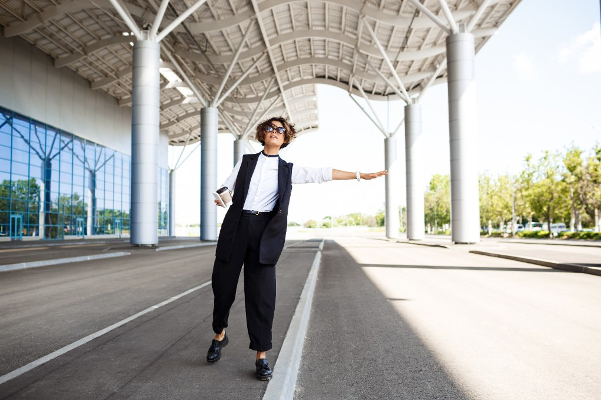 A woman in a dark suit outside a glass building, one arm out to hail a car.